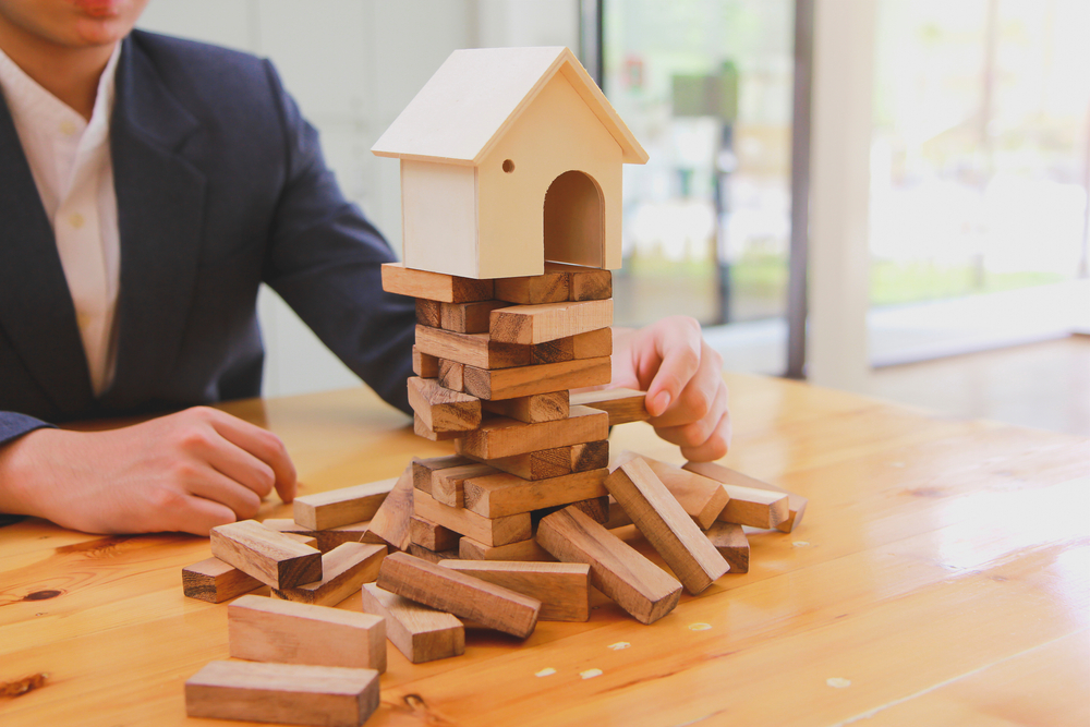 Real estate agent sitting at a table with a home on top of a Jenga game. Concept representing the pieces that threat the sale of a home on Long Island, including a home inspection.