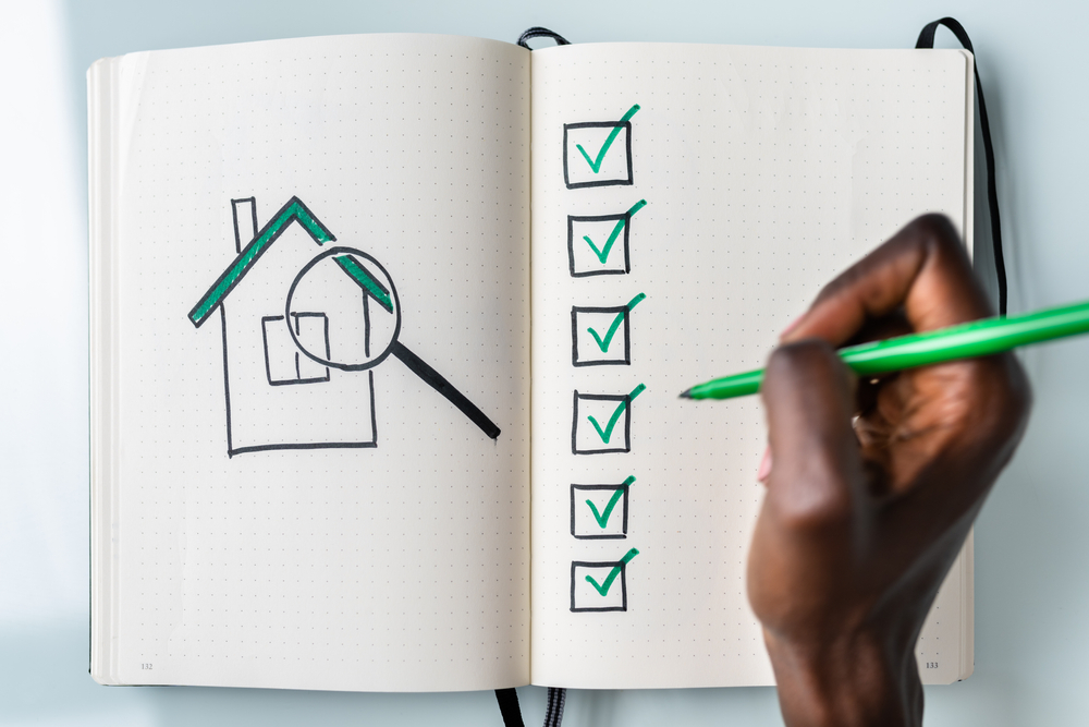 Man holding a pen, checking off a list of to-do items. Left side of the page has a house and magnifying glass drawn in pen. Concept of home buying checklist