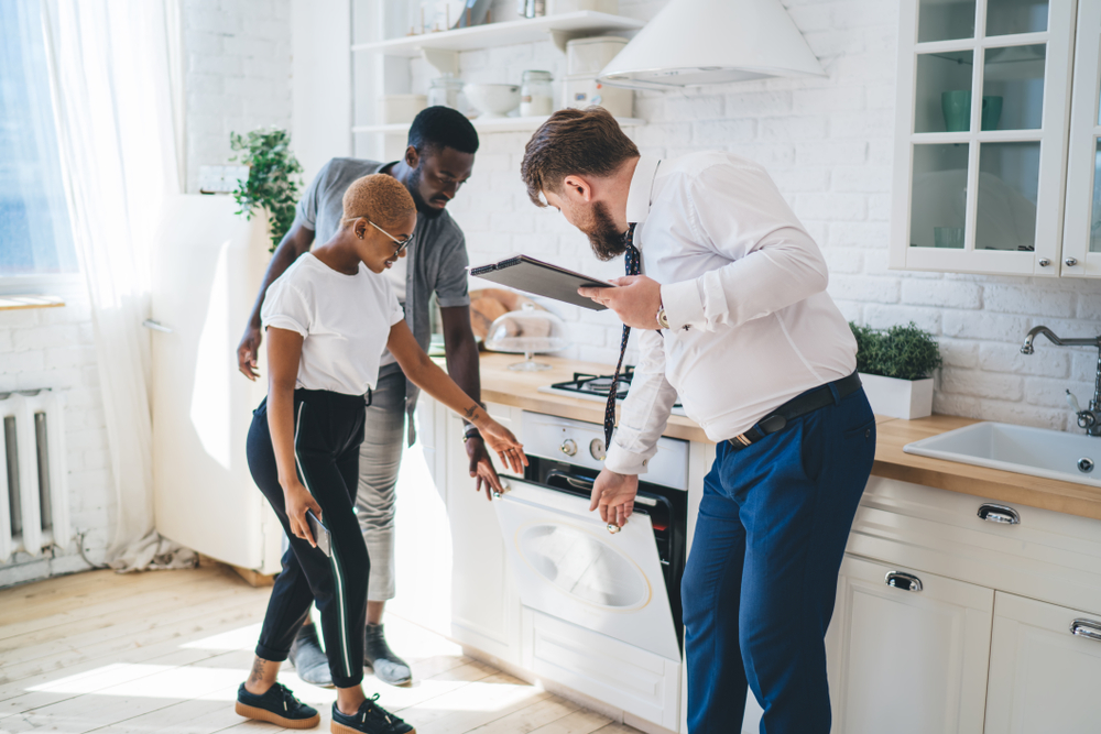 Real estate agent with clipboard looking in dishwasher and going over home inspection with a couple ready to buy a home